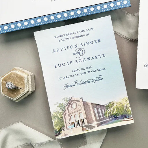 Save the Date with a church illustration, ring, and ribbon on a gray surface.