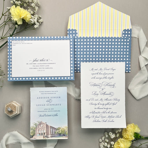 Wedding invitation set with blue and white polka dot patterned envelope on a gray surface with yellow flowers.