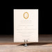 Charmain Invitation card with gold emblem on a stand against a black background