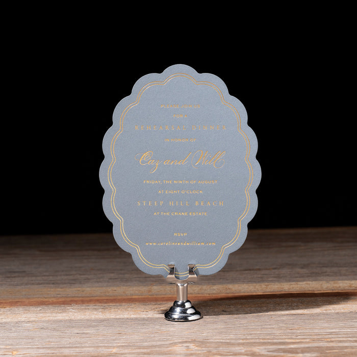 Scalloped Oval Rehearsal Dinner card on a stand with a dark background