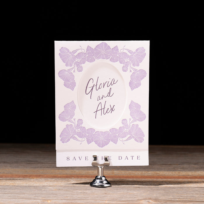 Gloria Save the date card with floral design and text on a stand against a black background