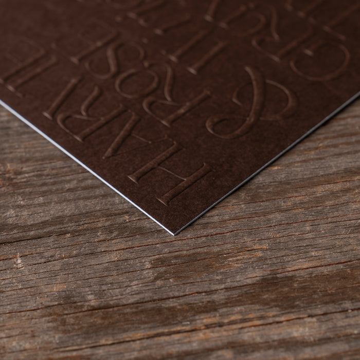 Edgepainted corner detail of dark brown embossed text invitation on a wooden surface