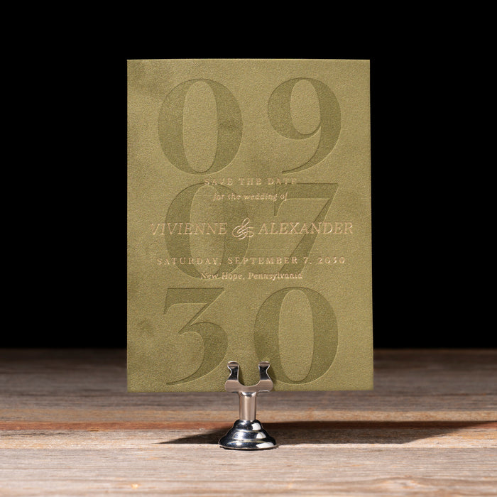 Green save the date card with debossed numbers on a stand against a dark background