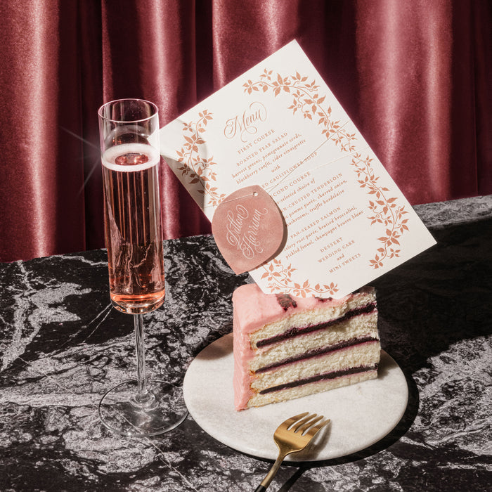 Elaborate wedding menu on slice of layered cake with a glass of pink champagne on a marble surface, against a red curtain background.