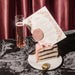 Elaborate wedding menu on slice of layered cake with a glass of pink champagne on a marble surface, against a red curtain background.