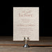 Celina rehearsal dinner card with intricate text on a wooden stand against a dark background