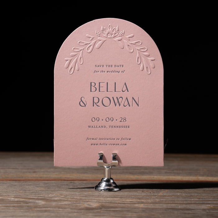 Pink save the date card with floral design on a wooden stand against a black background
