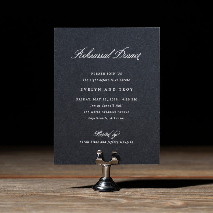 Dark blue rehearsal dinner card on a stand with a dark background