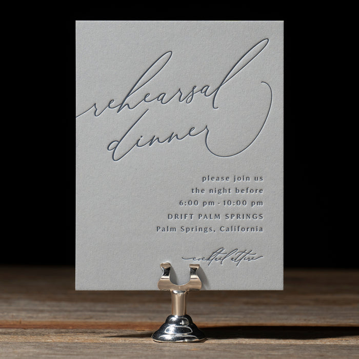 Gray blue rehearsal dinner invitation card on a stand with a black background