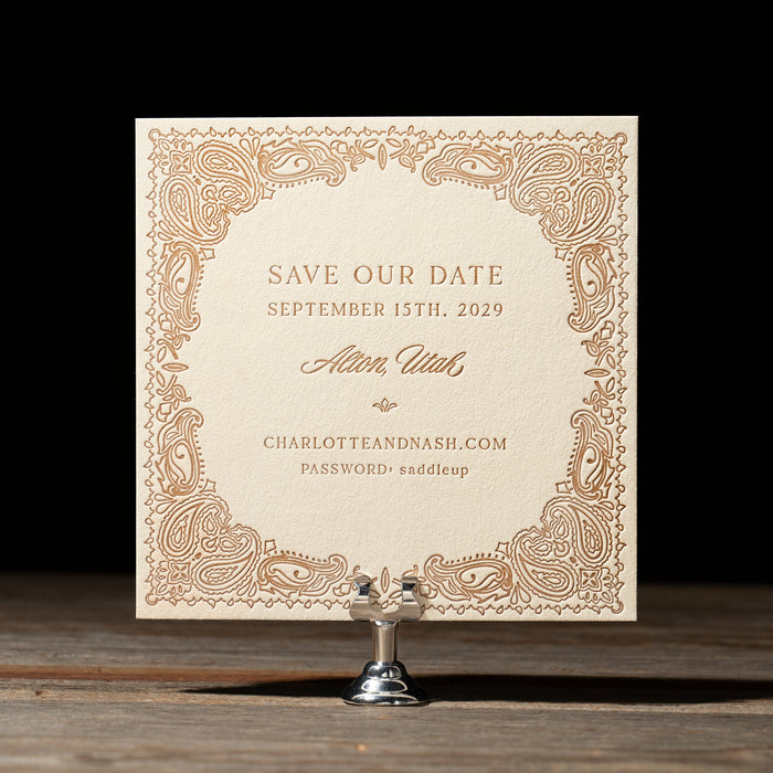 Decorative Nash 'Save the Date' card with intricate patterns on a wooden stand against a black background.