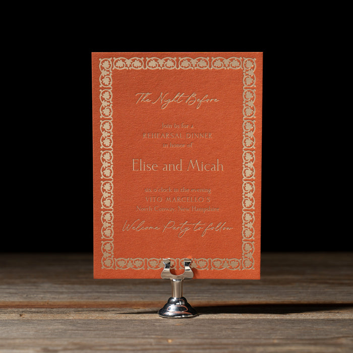 Rehearsal Dinner card with decorative border on a stand on a wooden surface