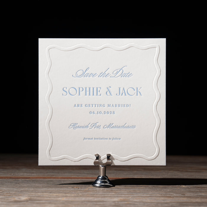 Ryann 'Save the Date' card with blue text on a wooden stand against a black background