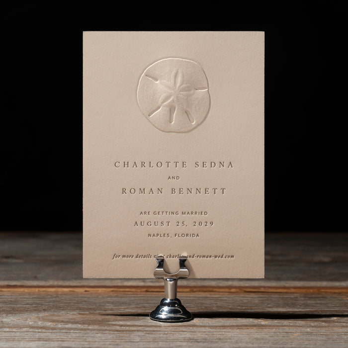 Beige Sedna save the date card on a wooden stand with a dark background