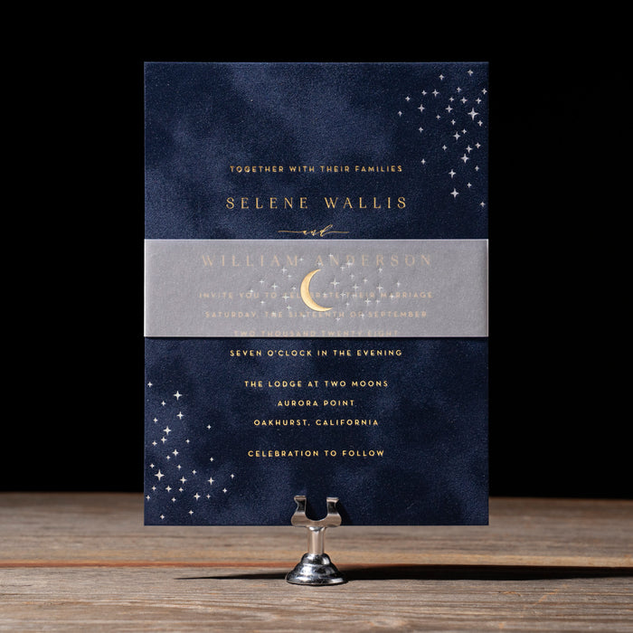 Navy blue wedding invitation with gold text on a wooden stand against a black background