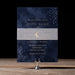 Navy blue wedding invitation with gold text on a wooden stand against a black background
