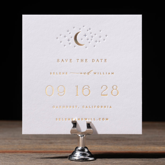 Selene 'Save the Date' card with gold text on a wooden stand against a black background