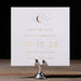 Selene 'Save the Date' card with gold text on a wooden stand against a black background