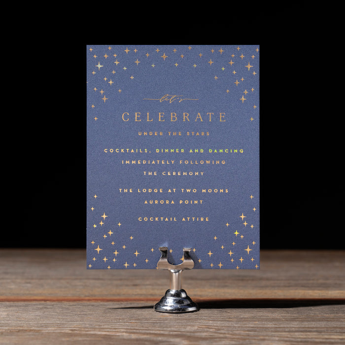 Navy blue event invitation with gold text and star patterns on a wooden stand against a dark background