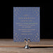 Navy blue event invitation with gold text and star patterns on a wooden stand against a dark background